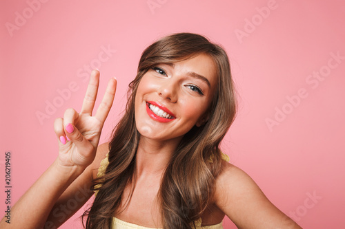 Photo of cheerful pretty woman 20s with long brown hair smiling and showing victory sign at camera while taking selfie on mobile phone, isolated over pink background