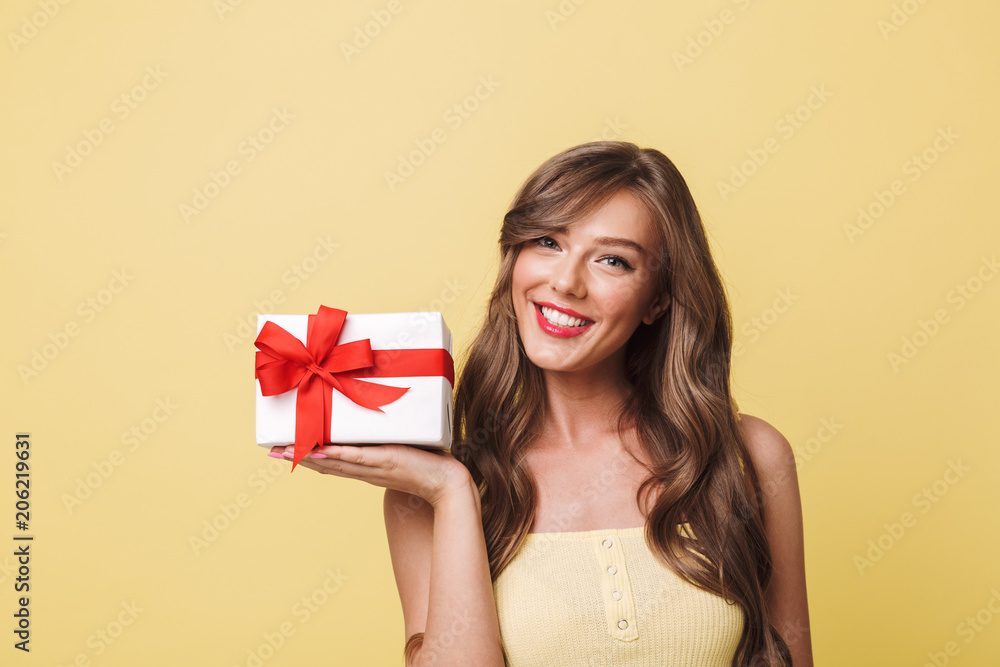 Photo of fascinating woman 20s having long brown hair smiling and holding present box wrapped with bow at her palm, isolated over yellow background