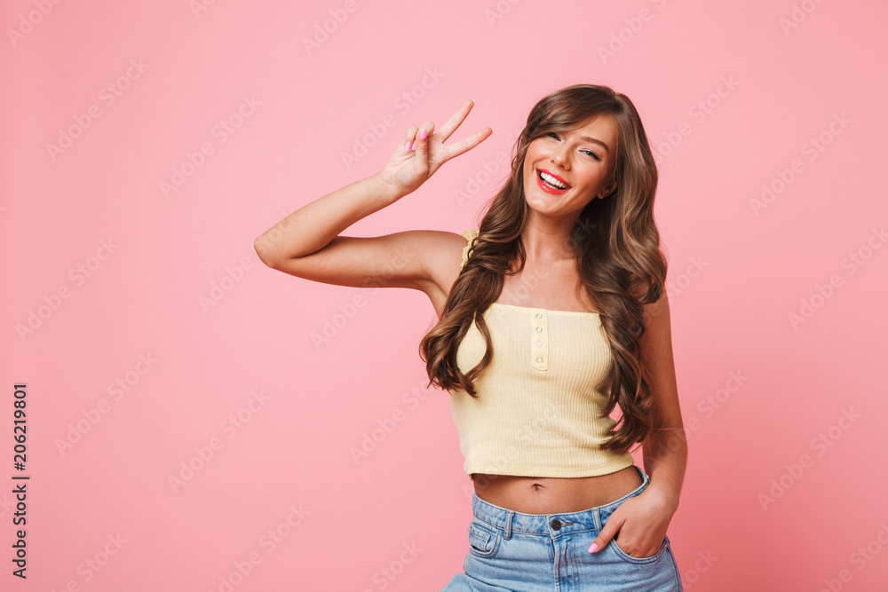 Photo of happy vacation girl 20s with long brown hair wearing summer clothing smiling and showing peace sign at camera, isolated over pink background Photo of happy vacation girl 20s with long brown hair wearing summer clothing smiling and showing peace sign at camera, isolated over pink background