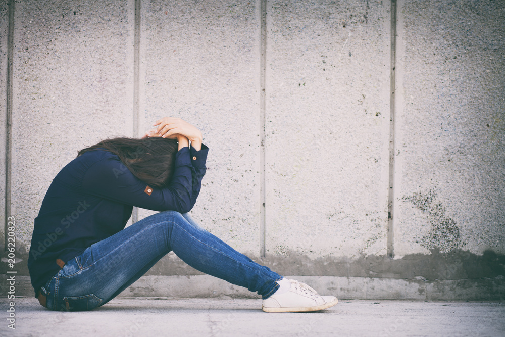 A teenage girl with depression sits alone on a concrete wall background