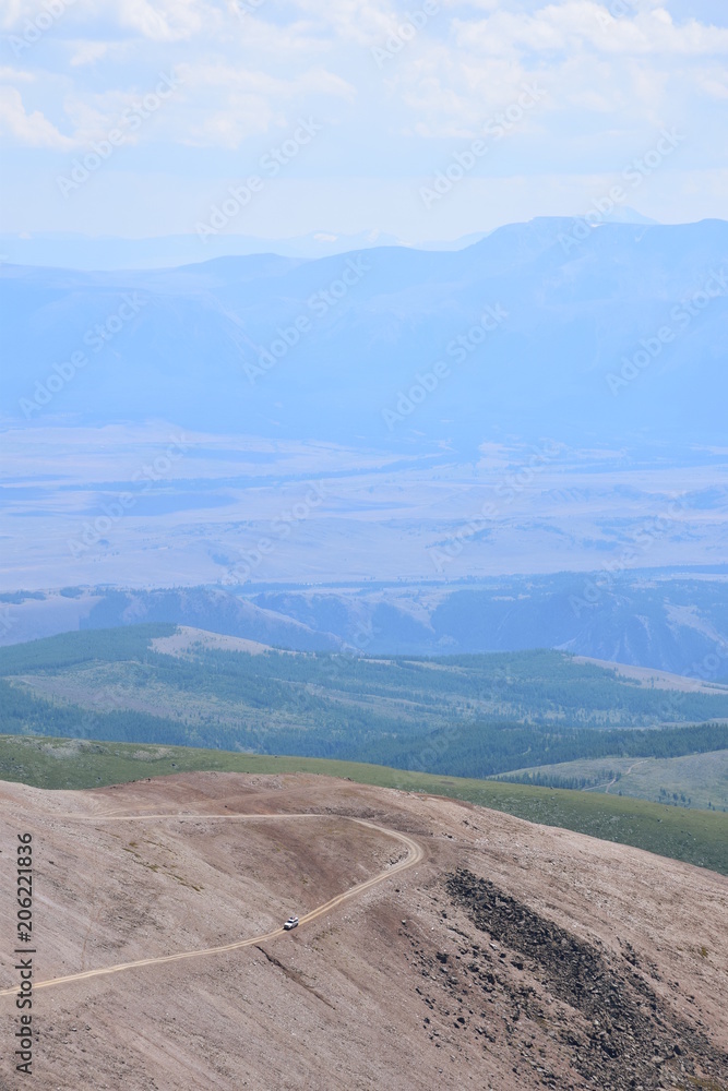 An elevated view of a car driving over a mountain dirt road with a vast valley in the background