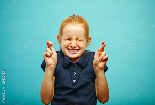 Children girl tightly closed his eyes and put fingers crossed, make a wish, believe in the dream, expresses heartfelt emotions, has funny facial features, beautiful red hair with freckles. photo