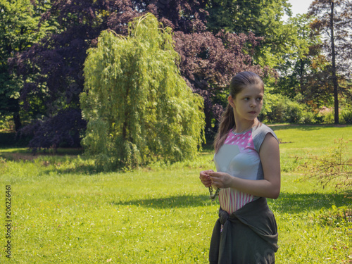 Sad lonely caucasian teenage girl stands half-turned to the camera holding a wreath of flowers, and looks outside the frame,with the green summer lawn of the park surrounded by trees on the background photo