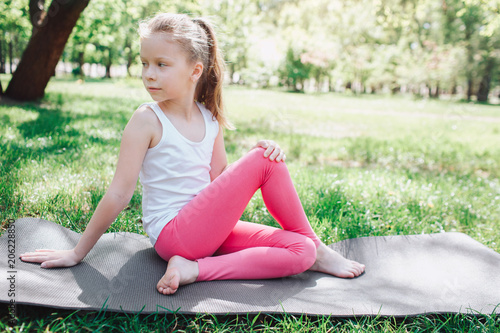 Cool girl in pink pants is sitting on carimate in park and looking back. She is keeping her legs crossed. She is leaning on her hands. Yoga and Pilates Concept