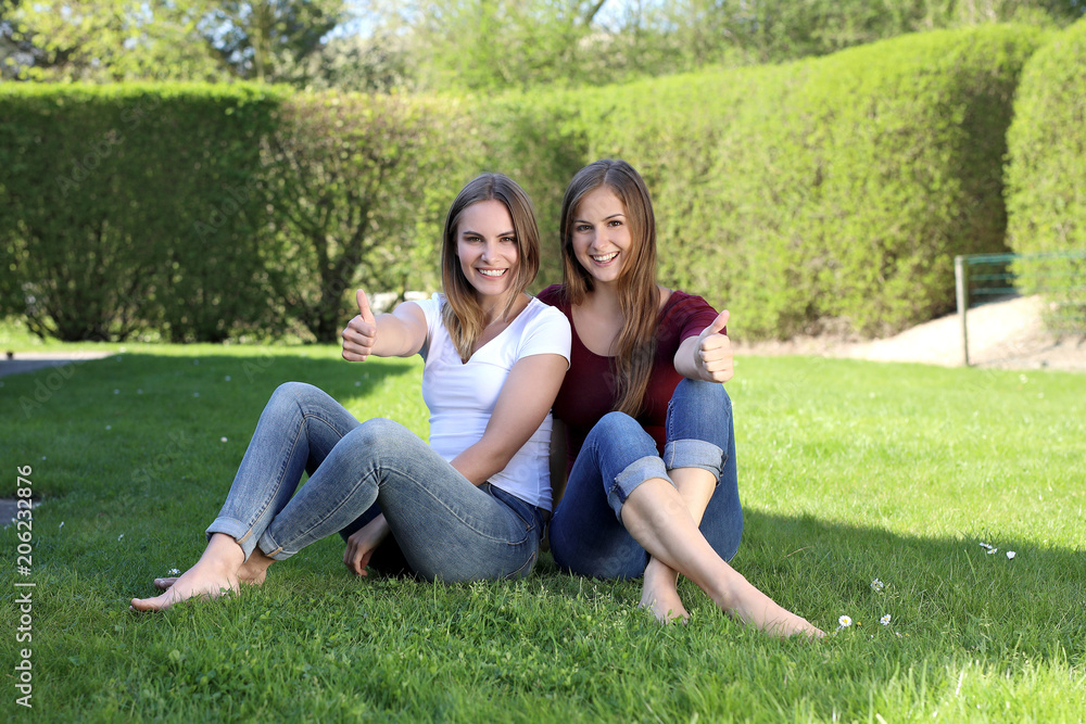 Zwei hübsche blonde Frauen mit nackten Füßen sitzen lachend auf einem Rasen und zeigen Daumen hoch