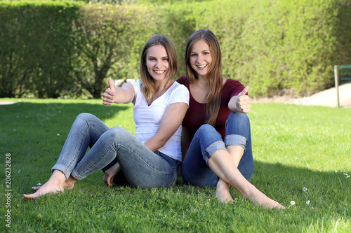 Zwei hübsche blonde Frauen sitzen lachend im Gras und zeigen Daumen hoch