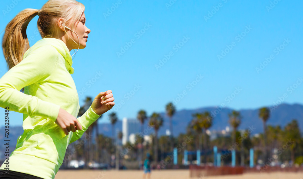 fitness, sport and technology concept - happy woman running and listening to music in earphones over venice beach background in california