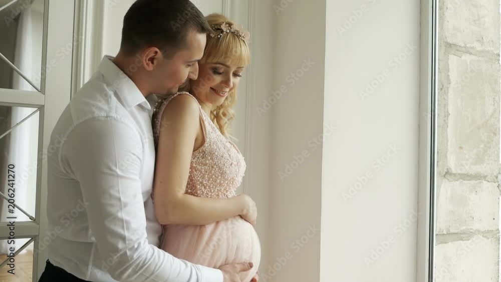 man husband embracing belly of his pregnant wife in dress standing next to window man husband embracing belly of his pregnant wife in dress standing next to window