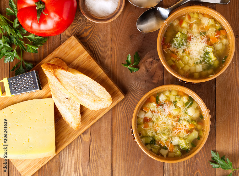 Two bowl of minestrone soup with cheese on cutting board and vegetables on rustic wooden background, top view.