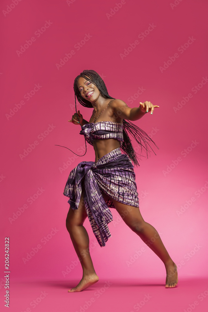 Beautiful African Black girl wearing traditional colorful African outfit does a dramatic dance move against a colorful pink background