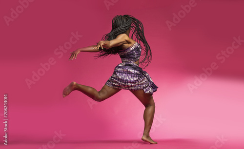 Fototapeta Naklejka Na Ścianę i Meble -  Beautiful African Black girl wearing traditional colorful African outfit does a dramatic dance move against a colorful pink background