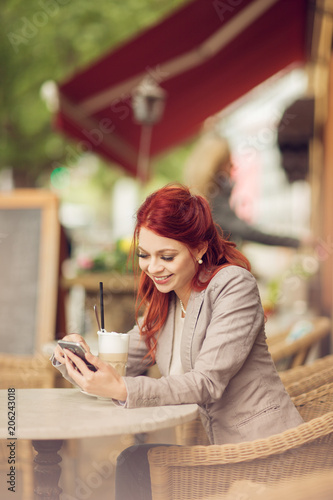 young beautiful woman in a street cafe enjoying a coffee, is busy with her cell phone, summerly urban mood