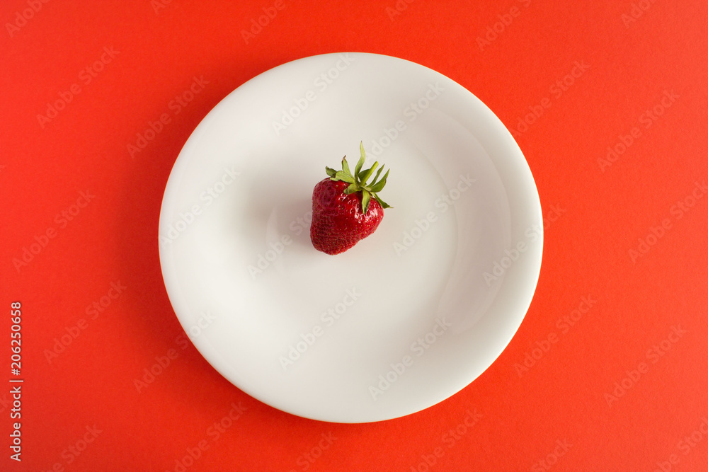 One strawberry in the white plate on the red background.Top view.Diet minimal concept. One strawberry in the white plate on the red background.Top view.Diet minimal concept.