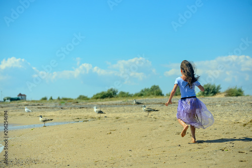 Happy little girl plays and runs along the beach on a Sunny day in a purple dress, and disperses seagulls on the sand. The horizontal orientation of the image