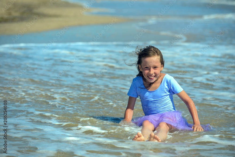 Happy little girl in purple dress sitting in the water on the beach on a Sunny day, looking at the camera and laughing. Horizontal orientation Happy little girl in purple dress sitting in the water on the beach on a Sunny day, looking at the camera and laughing. Horizontal orientation