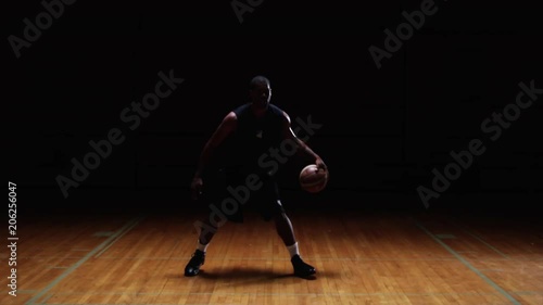 A basketball player dribbles the ball through his legs on a dark court