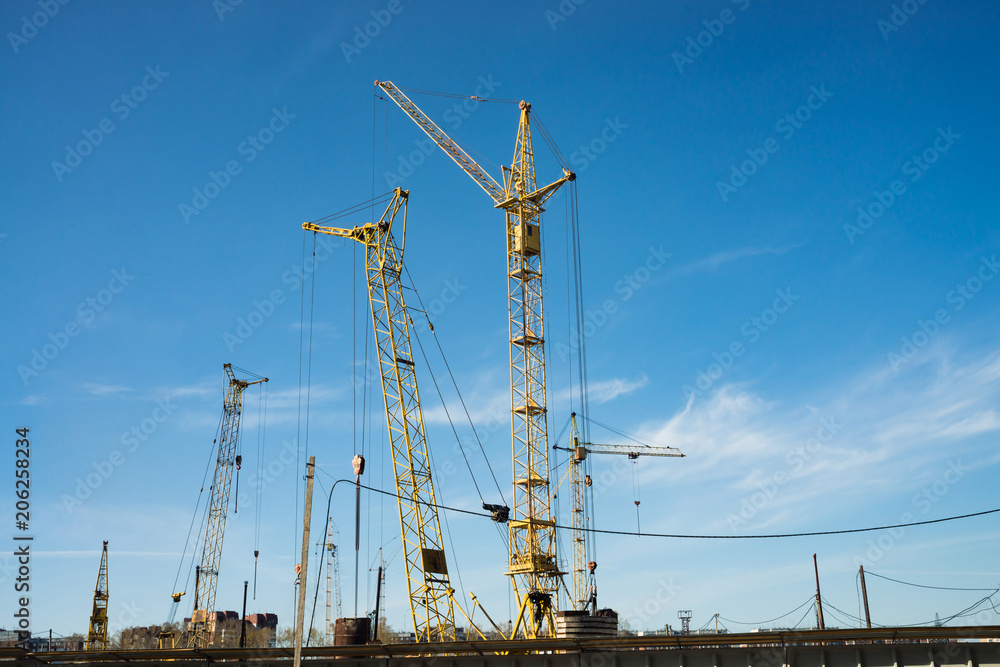 Big tower cranes above buildings under construction against blue sky. Background image of construction close-up with copy space. Build of city. Big tower cranes above buildings under construction against blue sky. Background image of construction close-up with copy space. Build of city.