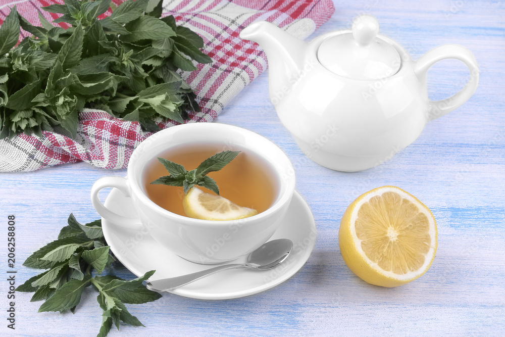 White cup of tea with mint and lemon and a kettle on a blue wooden background White cup of tea with mint and lemon and a kettle on a blue wooden background