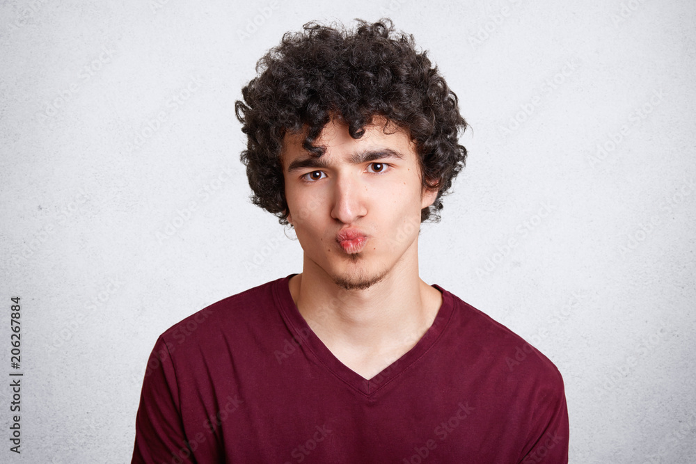 Attractive young male with crisp dark hair keeps mouth round, makes grimace, wears casual sweater, looks directly into camera, isolated over white studio concrete wall. People, youth concept Attractive young male with crisp dark hair keeps mouth round, makes grimace, wears casual sweater, looks directly into camera, isolated over white studio concrete wall. People, youth concept