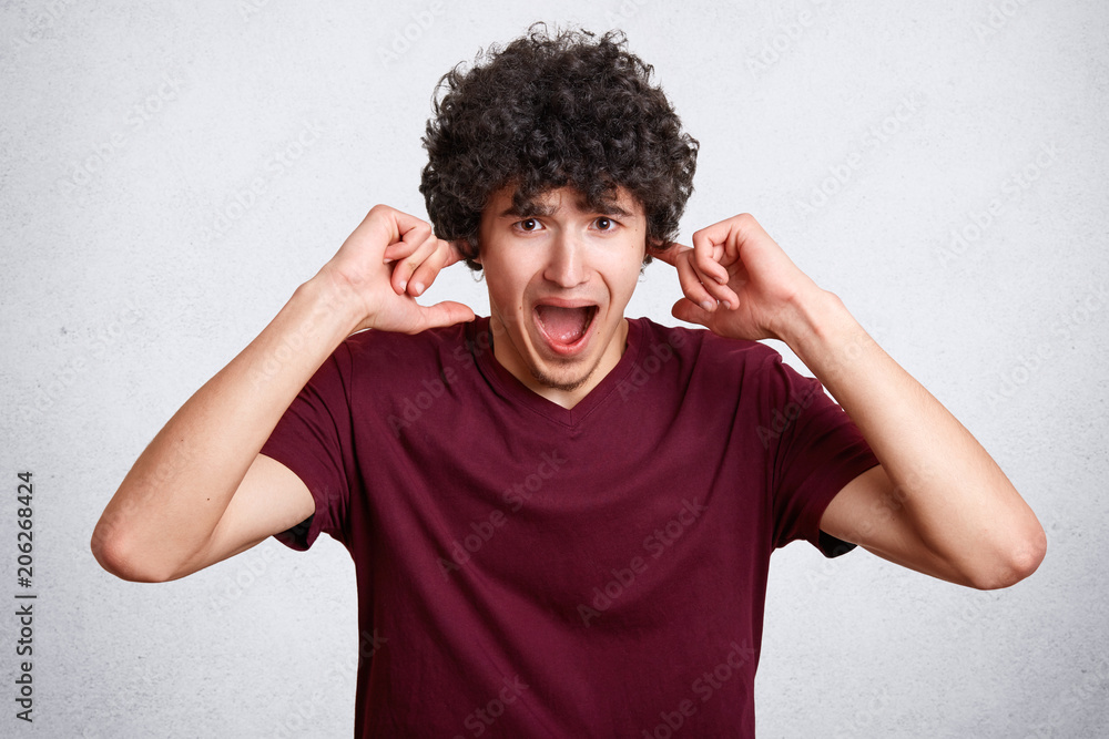 Stressful curly male plugs ears, being in panic, avoids loud irritating sound, wears casual t shirt, stands against white background. Annoyed handsome teenager doesn`t want to hear pop music. Stressful curly male plugs ears, being in panic, avoids loud irritating sound, wears casual t shirt, stands against white background. Annoyed handsome teenager doesn`t want to hear pop music.