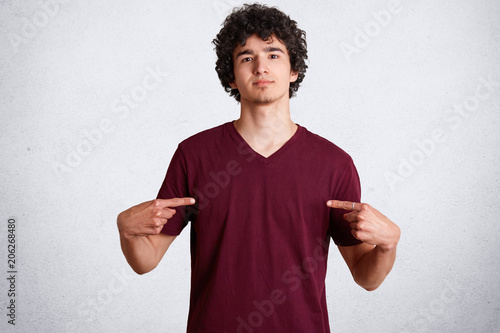 Portrait of handsome young male indicates at blank space of casual t shirt for your advertisement or designing content, has crisp hair, poses against white concrete wall. Look at my new clothes.