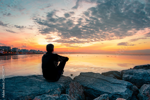Fotografie hombre joven sentado en una roca mirando el amanecer delante del mar