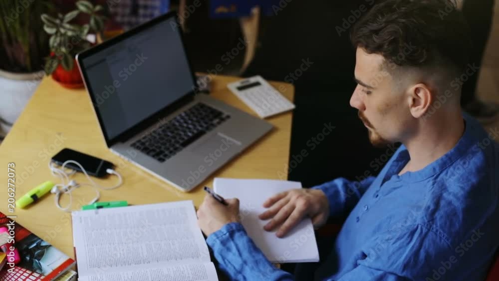 top view of handsome bearded male student noting text from education book notice copy writing study process man sitting at table working with laptop notebook studing home interior workplace indoors top view of handsome bearded male student noting text from education book notice copy writing study process man sitting at table working with laptop notebook studing home interior workplace indoors