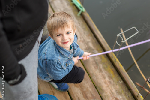 Portrait of cute little blond boy sitting on wooden pier near father. Adorable child having fun and smiling at lake or river shore. Father protection and care, happy childhood concept