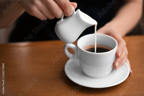 Woman pouring milk into cup of coffee on table