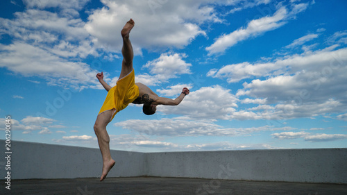 Tricking on street. Martial arts. Man flips back barefoot. Shooted from bottom foreshortening against sky.