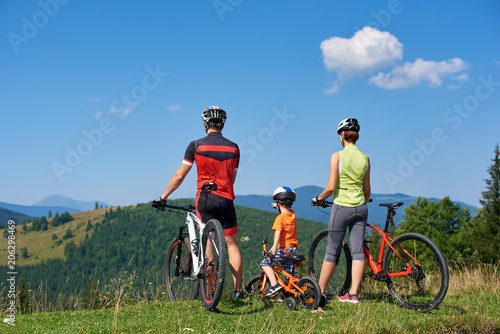 Fototapeta Naklejka Na Ścianę i Meble -  Back view of sportsman family cyclists, mother, father and kid resting with bikes on top of grassy hill, enjoying beautiful mountains view. Child pointing into a distance. Active lifestyle concept