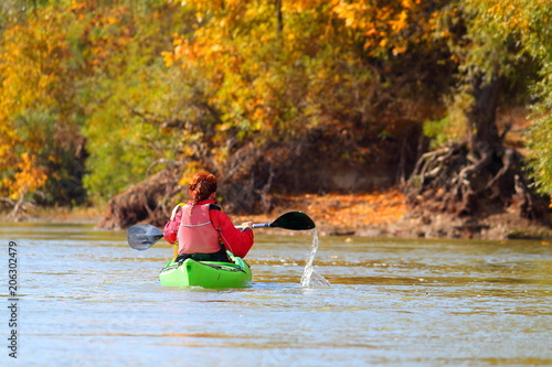 A girl in a green kayak and a red lifejacket is rowing along the Danube River against the background of autumn yellow trees. Autumn kayaking. The concept of a healthy lifestyle