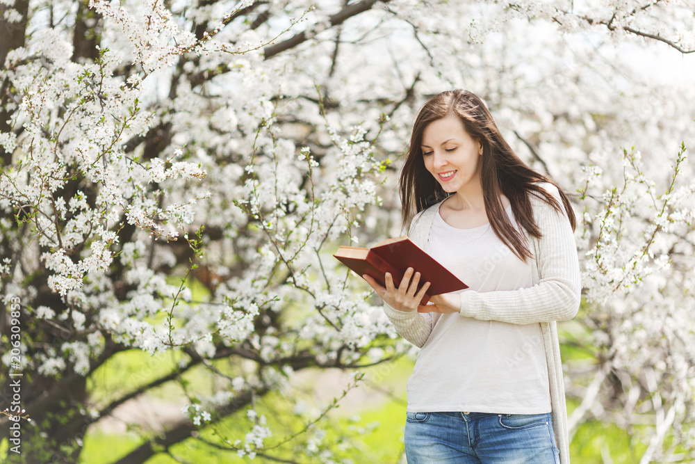 Young smiling relaxed beautiful woman in light casual clothes reading interesting book standing in city garden or park on blooming tree background. Spring nature, flowers. Lifestyle, leisure concept. Young smiling relaxed beautiful woman in light casual clothes reading interesting book standing in city garden or park on blooming tree background. Spring nature, flowers. Lifestyle, leisure concept.