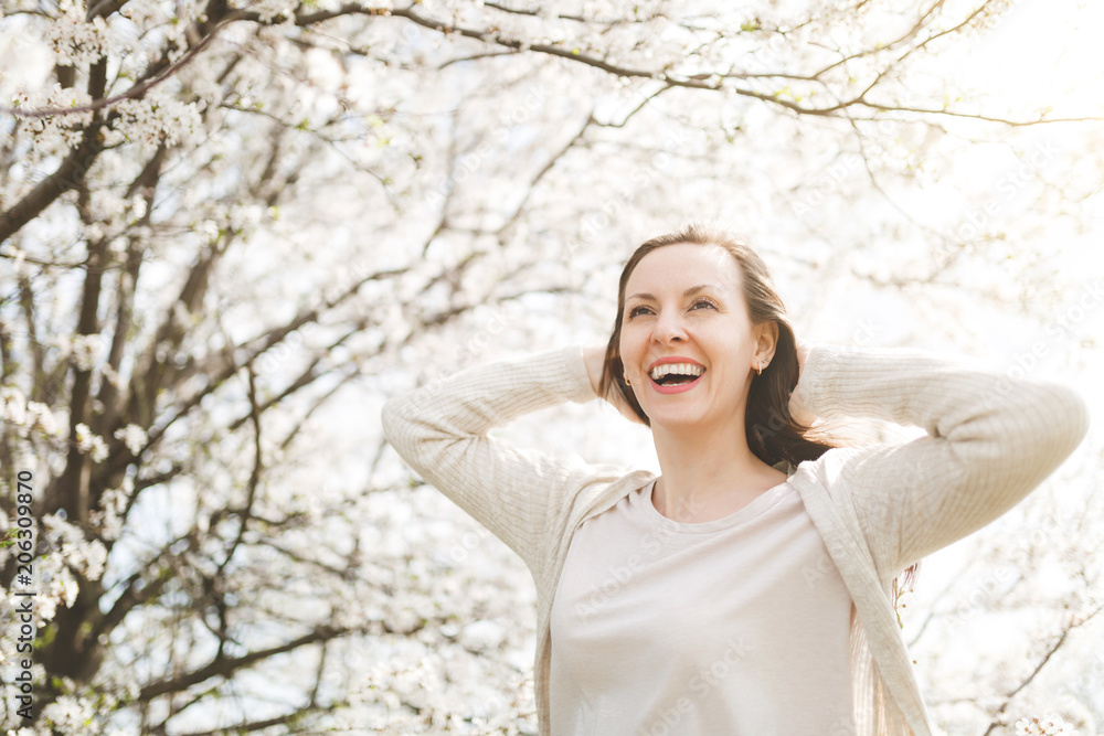 Young relaxed happy attractive woman in light casual clothes kepping hand behind head standing in city garden or park on blooming tree background. Spring nature, flowers. Lifestyle, leisure concept. Young relaxed happy attractive woman in light casual clothes kepping hand behind head standing in city garden or park on blooming tree background. Spring nature, flowers. Lifestyle, leisure concept.