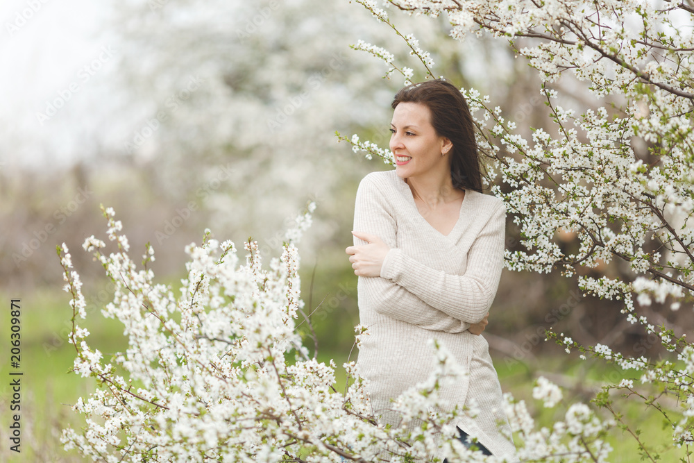 Young smiling happy charming woman in light casual clothes holding hands folded standing in city garden or park on blooming tree background. Spring nature, flowers. Lifestyle, leisure concept.