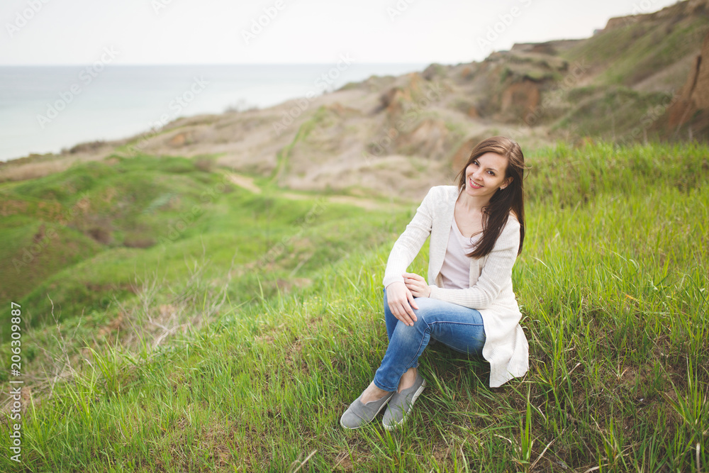 Young brunette smiling beautiful woman in light casual clothes sitting on grass with crossed hands and legs near water on green field background. Beautiful landscape. Lifestyle, leisure concept. Young brunette smiling beautiful woman in light casual clothes sitting on grass with crossed hands and legs near water on green field background. Beautiful landscape. Lifestyle, leisure concept.