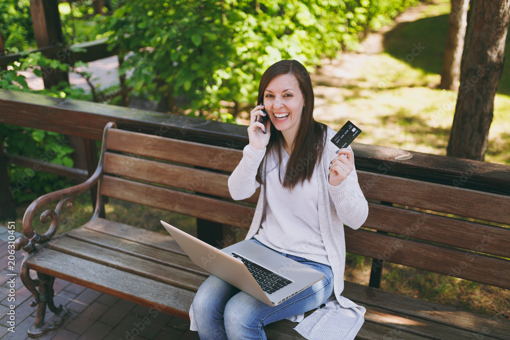 Female holding credit card, talking on mobile phone, having good pleasure conversation. Woman sitting on bench working on modern laptop in street outdoors. Mobile Office. Freelance business concept. Female holding credit card, talking on mobile phone, having good pleasure conversation. Woman sitting on bench working on modern laptop in street outdoors. Mobile Office. Freelance business concept.