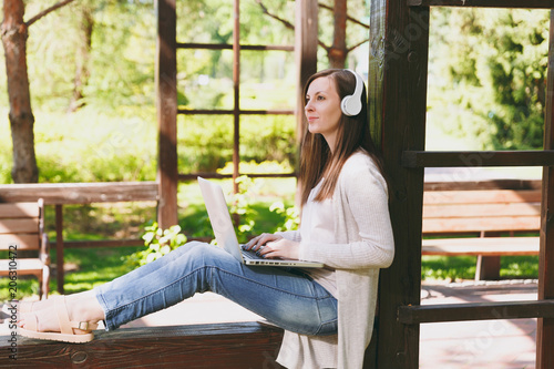 Free Download Young businesswoman in light casual clothes. Woman working on modern laptop pc computer, listen music in headphones on head in street outdoors. Mobile Office. Freelance business concept. Side view. - GFXLoad