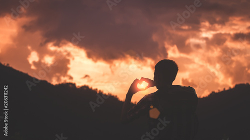 Young traveler man with backpack and standing at sunset and making by hands in shape of love heart.