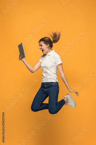 Image of young woman over orange background using laptop computer or tablet gadget while jumping.