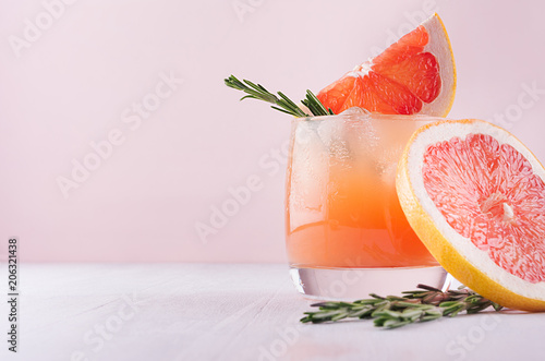 Cold lemonade of fresh grapefruit juice, alcohol and ice cubes decorated slice citrus and twig rosemary on fashion pastel pink background.