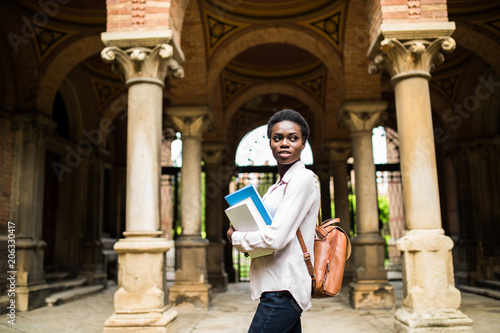 Portrait of a cheerful smiling african student girl wearing backpack and holding books at university campus