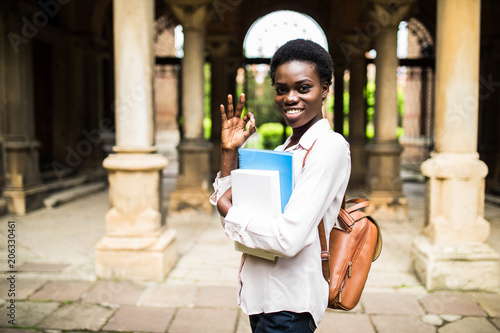Portrait of a cheerful smiling african student girl waving and wearing backpack and holding books at university campus