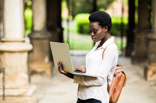 Portrait of smiling African american student doing homework outdoors using laptop for research. Positive dark skinned girl styling in university campus preparing for examinations outdoors photo