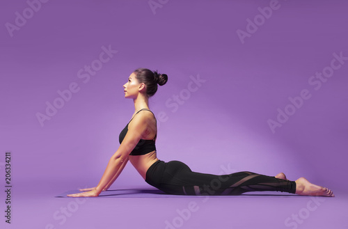 Long haired beautiful pilates or yoga athlete does a graceful pose while wearing a tight sports outfit against a bright purple background in a studio