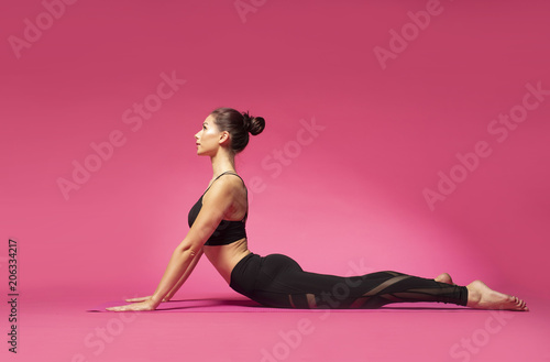 Long haired beautiful pilates or yoga athlete does a graceful pose while wearing a tight sports outfit against a pink background in a studio