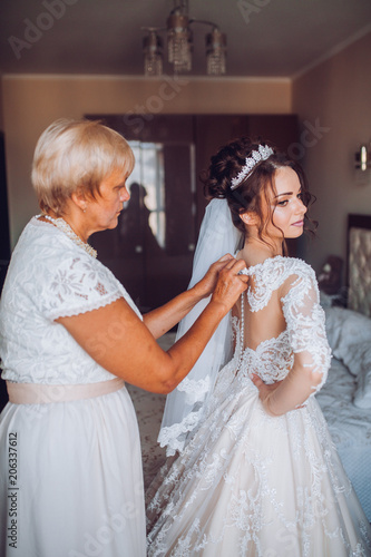 Beautiful bridesmaid and mother help put on wedding dress on gorgeous bride in luxury hotel room morning before the wedding