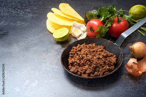 ingredients for tacos with roasted beef in a pan, tomatoes, onion, garlic, lime, avocado and herbs on a dark stone background with copy space