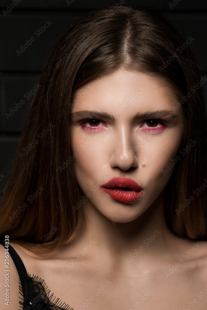 Close-up portrait of a beautiful fashion girl with big red lips on the background of a black brick wall, in a black sexy singlet. Biting his lip. Look. Bright sexy makeup. Close-up portrait of a beautiful fashion girl with big red lips on the background of a black brick wall, in a black sexy singlet. Biting his lip. Look. Bright sexy makeup.