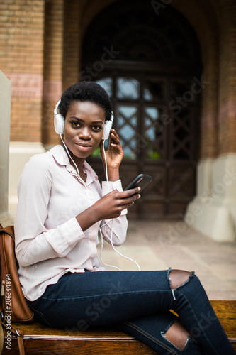 Beautiful afro american girl listening to music through headphones sitting on a bench. female with afro hair watching video on smartphone relaxing outside photo